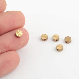 May include: Small, round, brass beads with a faceted top and a small hole. One bead rests on a finger, while four others are scattered on a white surface. These beads are ideal for jewelry making and crafting projects.