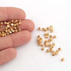 Puede incluir: Un montón de cuentas de metal de color dorado, algunas están en una mano, otras están esparcidas sobre una superficie blanca.
