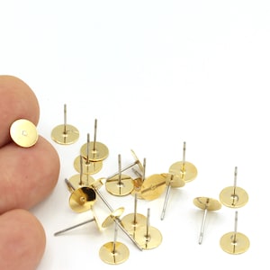 May include: A close-up of a pile of gold-colored earring posts with flat round backs. The posts are scattered on a white surface.