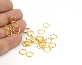May include: A collection of small, gold-colored jump rings, scattered on a white surface. Some rings are grouped together, while others are spread out. The rings appear to be made of metal and are open, ready for use in jewelry making or crafts.