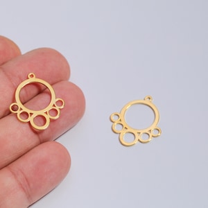 May include: Two gold-colored metal jewelry components. Each component features a large central circle with smaller circles attached. One is held in a hand, the other rests on a light gray surface. These components are designed for jewelry making.