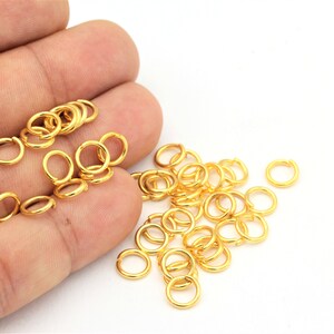 May include: A pile of gold jump rings, a type of jewellery finding, are scattered on a white surface. The rings are small and round, and they are perfect for connecting different pieces of jewellery together.