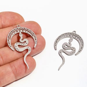 Puede incluir: Dos colgantes de metal plateado. Cada colgante presenta un diseño de serpiente dentro de una forma de media luna. La serpiente tiene un patrón detallado y la luna tiene un borde decorativo. Los colgantes se muestran sobre un fondo blanco.