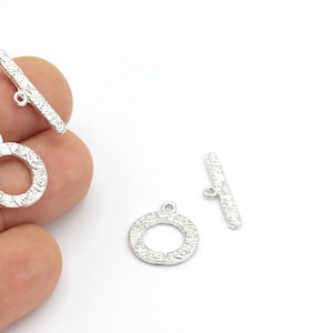 May include: Two silver-toned toggle clasps with a decorative, ornate design. Each clasp features a round ring and a straight bar.