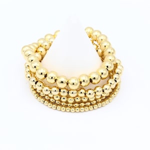 May include: A set of four gold beaded bracelets, each with a different size bead. The bracelets are stacked on top of each other, creating a layered look.