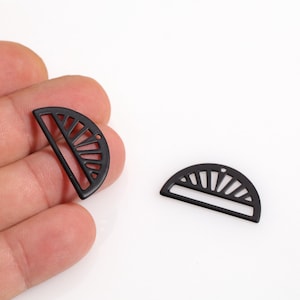 May include: Two black metal half-circle charms with a sunburst design. One charm is held in a hand, while the other lies on a white surface. These charms are suitable for jewelry making or craft projects.