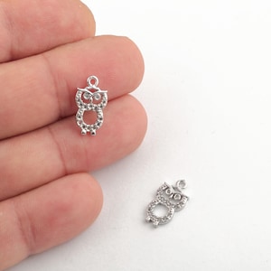 Op de afbeelding: Twee kleine, zilverkleurige uilenbedels met heldere kristallen accenten. De bedels hebben de vorm van een uil met een lus aan de bovenkant om aan sieraden of andere knutselwerken te bevestigen. Eén bedel wordt in een hand gehouden.