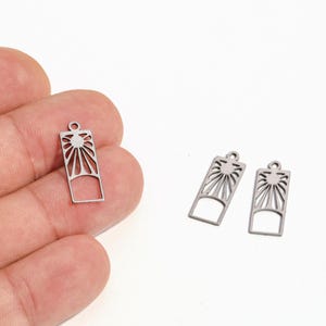 May include: Silver-colored metal charm pendants with a sunburst design. The pendants are rectangular with a small loop at the top for attaching to jewelry. One pendant is held in a hand, and two others are on a white background.