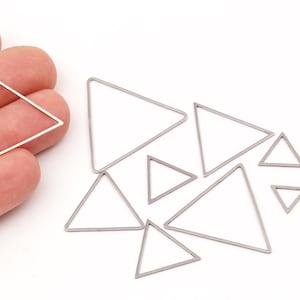 Puede incluir: Una colección de dijes triangulares plateados de marco abierto en varios tamaños. Un triángulo se sostiene en una mano, mientras que otros están dispersos sobre una superficie blanca. Estas formas geométricas son ideales para la fabricación de joyas o proyectos de artesanía.