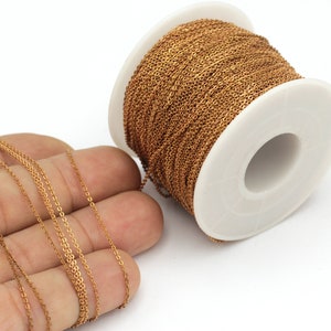 May include: A spool of gold-colored chain, approximately 10 feet long, is shown on a white surface. The chain is also shown laid out on a hand.