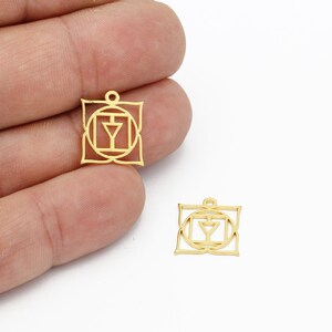 Puede incluir: Dos amuletos de metal de color dorado en forma de cuadrado con un símbolo en su interior. El símbolo es un círculo con un triángulo en su interior, rodeado de un cuadrado y un diseño floral.