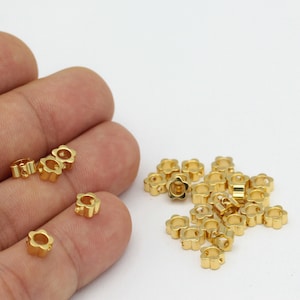 May include: A pile of gold-colored metal flower-shaped beads with a hole in the center. The beads are scattered on a white surface.