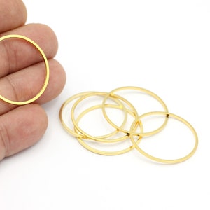 Può includere: Una collezione di anelli metallici circolari, sottili e color oro. Un anello è tenuto in mano, mentre altri sono sparsi su una superficie bianca. Questi anelli sono probabilmente componenti per la creazione di gioielli o progetti artigianali.
