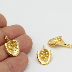 Puede incluir: Amuletos de metal dorado con un diseño en forma de escudo. Los amuletos tienen un lazo en la parte superior para sujetarlos a la joyería. El escudo tiene una superficie texturizada y un interior detallado con pequeños detalles redondos.