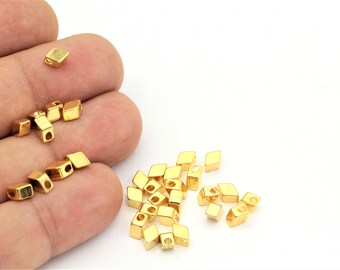 May include: A close-up of a pile of gold-colored metal beads. The beads are shaped like small diamonds with a hole in the center. The beads are scattered on a white surface.