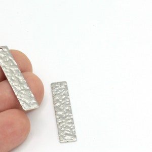 May include: Two silver metal rectangular pendant charms with a textured surface. The charms have a hole at the top for attaching to jewellery.