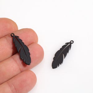 May include: Two black feather-shaped charms. One charm is held in a hand, the other is on a white surface. The charms have a small loop at the top for attaching to jewelry. The feathers have detailed veining.
