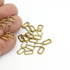 May include: A pile of gold-colored metal D-rings, also known as jump rings, are scattered on a white surface. Some of the rings are held in a hand.