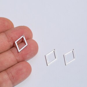 May include: Three small, silver-colored, open-frame diamond-shaped charms. One charm is held in a hand, while two others are on a white surface. The charms have a small hole at the top for stringing.