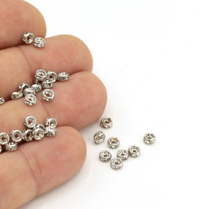 May include: A close-up of a hand holding a pile of silver-toned metal beads with clear rhinestones. The beads are scattered on a white surface.