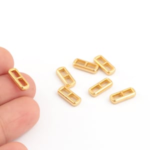 May include: Gold-coloured metal jump rings, also known as split rings, are scattered on a white surface. One ring is being held by a finger.