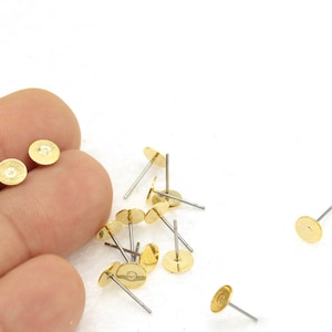 Könnte beinhalten: Nahaufnahme von goldfarbenen Ohrring-Findings mit einer flachen, runden Oberseite und einem silbernen Stift. Die Findings sind auf einer weißen Oberfläche verstreut.