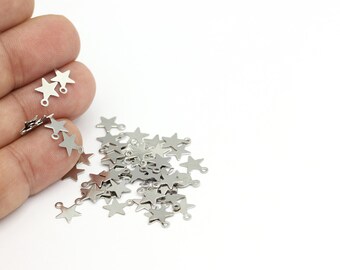 Puede incluir: Un montón de abalorios de estrella plateados con un anillo de salto en cada estrella. Los abalorios están esparcidos sobre una superficie blanca.