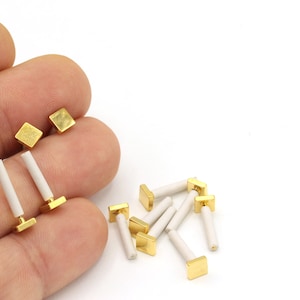 May include: A set of small, gold-colored square-shaped decorative pieces with white ceramic posts. The pieces are arranged on a white surface.