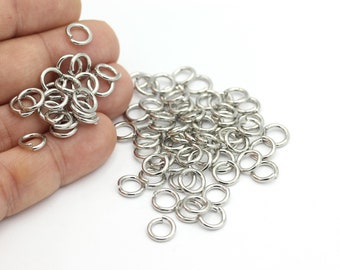Könnte beinhalten: Ein Haufen silberner Sprungringe, eine Art Schmuckzubehör, auf einer weißen Oberfläche. Die Ringe sind klein und rund und eignen sich perfekt zum Verbinden verschiedener Schmuckstücke.