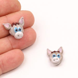 Może przedstawiać: Dwie szare szklane koraliki w kształcie głowy osła z niebieskimi oczami i brązowymi kokardkami. Koraliki mają około 2,5 cm wysokości.
