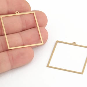 May include: Two gold-colored metal square frames with a small loop at the top. The frames are empty and ready to be filled with resin or other materials.
