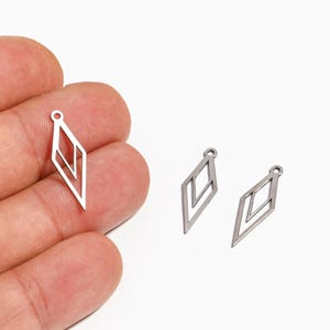 Op de afbeelding: Drie zilverkleurige, geometrische ruitvormige bedels. Eén bedel wordt in een hand gehouden, terwijl er twee andere op een wit oppervlak liggen. Elke bedel heeft een kleine lus aan de bovenkant om te bevestigen.