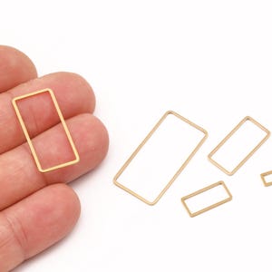 May include: Gold-colored rectangular metal jewelry findings in various sizes. One is held in a hand, with others scattered on a white surface. These open-frame rectangles are likely for crafting earrings, pendants, or other jewelry.