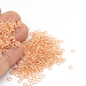 May include: A large pile of small, rose gold jump rings. The rings are scattered on a white surface.