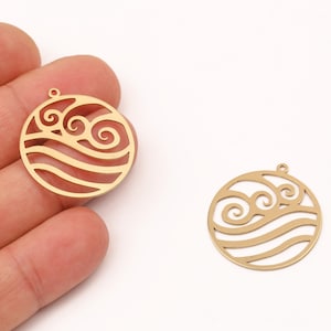 May include: Two gold-colored, circular pendants with a wave and swirl design. One pendant is held in a hand, while the other rests on a white surface. The pendants have a small loop at the top for attaching to a chain or other jewelry.