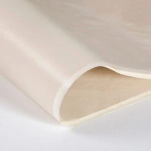 May include: A close-up of a sheet of off-white material, possibly fabric or paper, with a smooth, slightly reflective surface. The material is folded over, revealing a lighter interior and a rounded edge.
