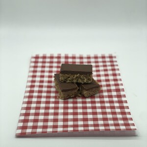 May include: Three rectangular dessert bars with a chocolate topping are stacked on a red and white checkered paper. The bars have a light brown, textured base. The paper has a classic picnic pattern.