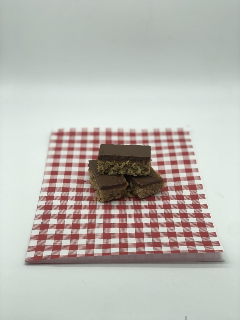 May include: Three rectangular dessert bars with a chocolate topping are stacked on a red and white checkered paper. The bars have a light brown, textured base. The paper has a classic picnic pattern.