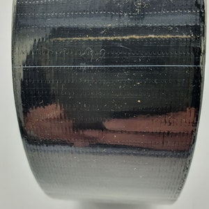 K&ouml;nnte beinhalten: Nahaufnahme einer Rolle schwarzes Klebeband mit strukturierter Oberfl&auml;che. Das Band ist eng gewickelt, mit einer d&uuml;nnen, transparenten Linie, die horizontal durch die Mitte verl&auml;uft. Die Oberfl&auml;che reflektiert Licht und zeigt Hinweise auf Braun und andere Farben.