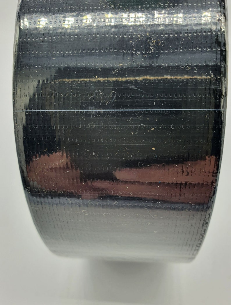K&ouml;nnte beinhalten: Nahaufnahme einer Rolle schwarzes Klebeband mit strukturierter Oberfl&auml;che. Das Band ist eng gewickelt, mit einer d&uuml;nnen, transparenten Linie, die horizontal durch die Mitte verl&auml;uft. Die Oberfl&auml;che reflektiert Licht und zeigt Hinweise auf Braun und andere Farben.