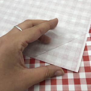 May include: A hand lifting a sheet of translucent paper from a red and white checkered surface. The paper is thin and partially see-through, revealing the checkered pattern beneath. The background features a similar red and white checkered pattern.