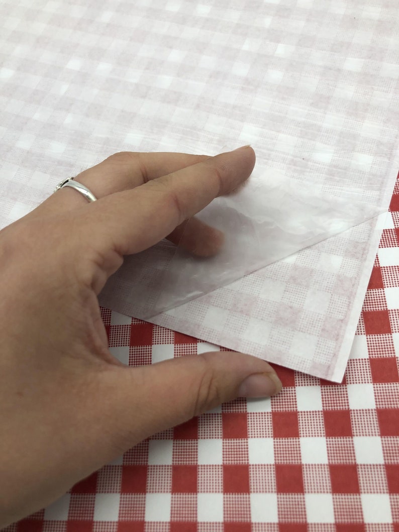 May include: A hand lifting a sheet of translucent paper from a red and white checkered surface. The paper is thin and partially see-through, revealing the checkered pattern beneath. The background features a similar red and white checkered pattern.