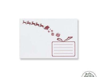 Könnte beinhalten: Weißer Umschlag mit einer weinroten Illustration von Santa Claus in seinem Schlitten, gezogen von Rentieren, die Geschenke liefern. Der Umschlag hat einen rechteckigen Bereich mit Linien für die Adresse des Empfängers und ein Schleifendesign.