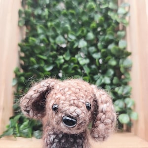 May include: A handmade, brown and gray crocheted dog with black eyes and a black nose. The dog has floppy ears and a fluffy texture. The background features green foliage and a wooden frame.