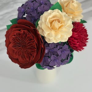 May include: A bouquet of paper flowers in a white vase. The arrangement includes a large burgundy rose, cream-colored roses, red and purple flowers, and green leaves. The flowers are arranged in a compact, full display.
