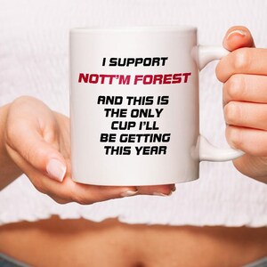 May include: White ceramic mug with the text "I SUPPORT NOTT'M FOREST" in black and red. The mug also has the text "AND THIS IS THE ONLY CUP I'LL BE GETTING THIS YEAR". The mug is held by a person.