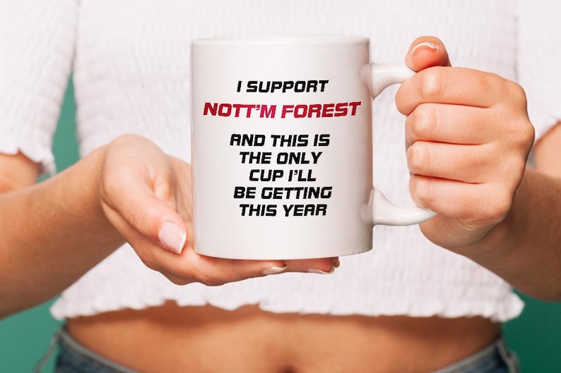 May include: White ceramic mug with the text "I SUPPORT NOTT'M FOREST" in black and red. The mug also has the text "AND THIS IS THE ONLY CUP I'LL BE GETTING THIS YEAR". The mug is held by a person.