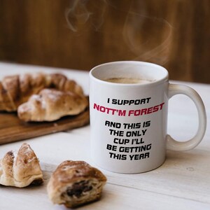 May include: White ceramic mug with the text "I SUPPORT NOTT'M FOREST" in black and red. The mug is filled with a hot beverage, and croissants are on a wooden board nearby.
