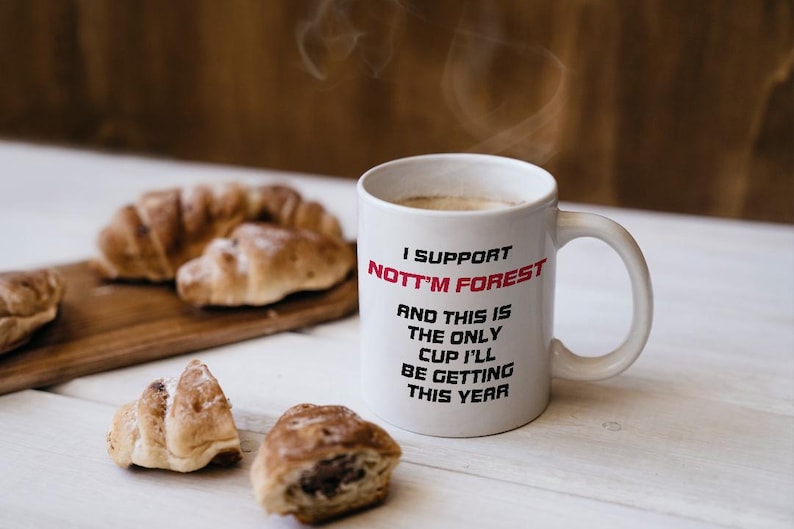 May include: White ceramic mug with the text "I SUPPORT NOTT'M FOREST" in black and red. The mug is filled with a hot beverage, and croissants are on a wooden board nearby.