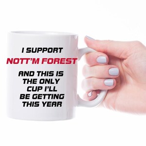 May include: White ceramic mug with the text "I SUPPORT NOTT'M FOREST" in black and red. The text continues with "AND THIS IS THE ONLY CUP I'LL BE GETTING THIS YEAR". The mug is held by a hand with light blue nail polish.